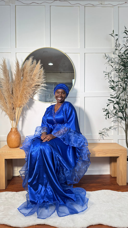 A woman wearing a vibrant royal blue traditional gown and matching headwrap sits on a light wood bench in front of a white paneled wall with a round mirror.